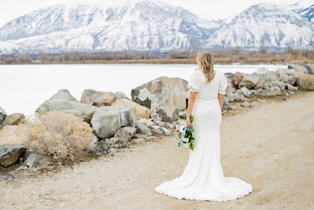 Bride standing with back to the camera and looking at the mountains in front of her. her bouquet is hanging next to her