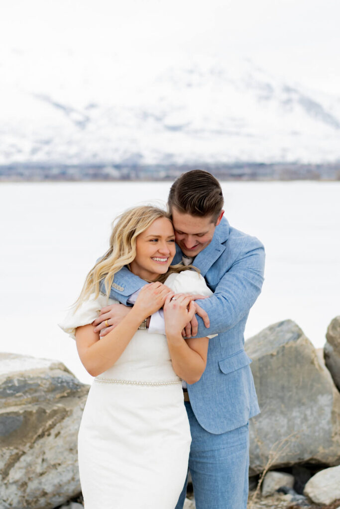 bride hugged from behind by groom with mountain and lake behind them
