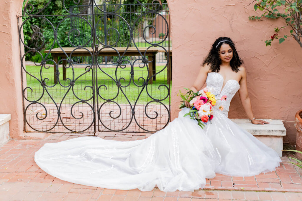 Bride sitting on a bench near a gate with her dress spread out.