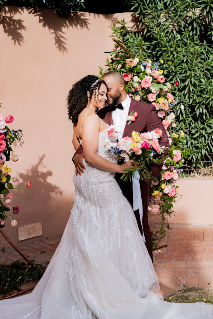 Bride and groom close together in front of a flower arch. Both are looking down.