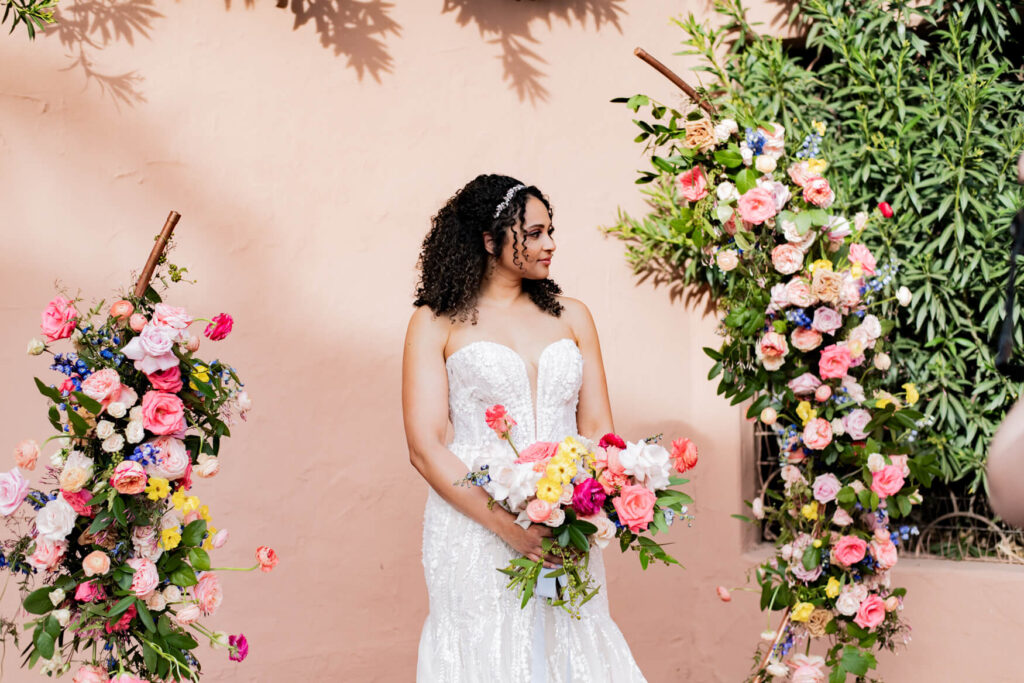 Bride standing in the middle of her flower arch while looking away and holding her bouquet.