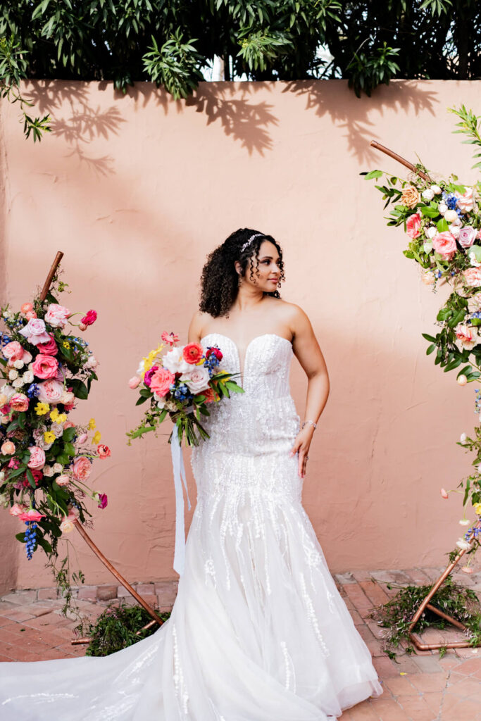 Bride standing in the middle of her flower arch while looking away and holding her bouquet.