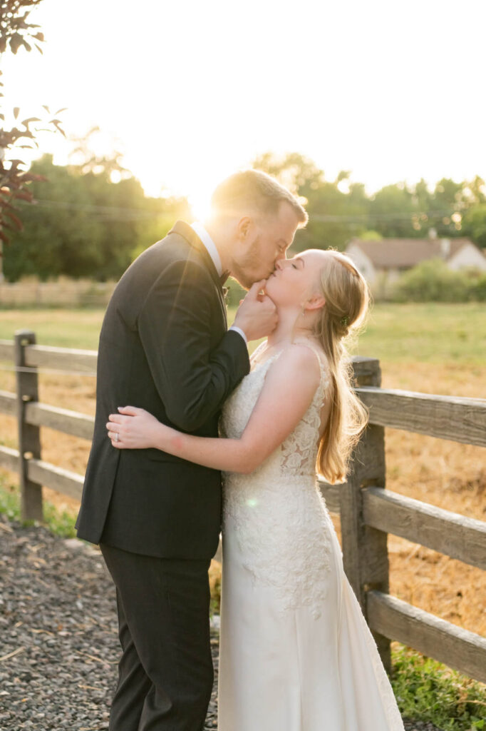 Bride and groom standing together and kissing against a fence with the sun shining from behind them