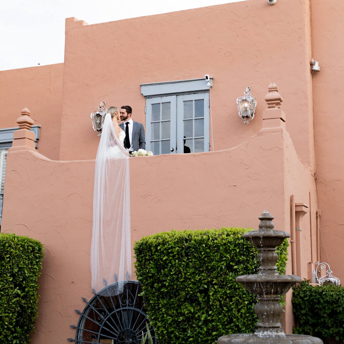 Bride and groom standing on a balcony and kissing with the bride's veil hanging off the balcony.