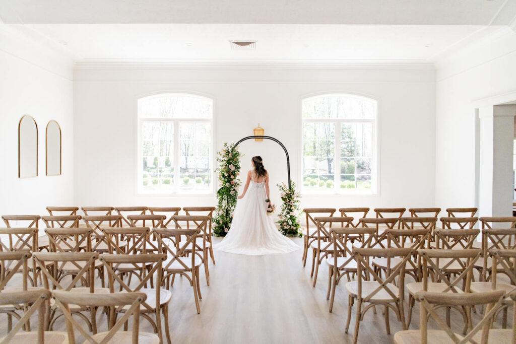 Bride standing with her back to the camera at the front of the ceremony space with her dress laid out behind her